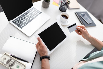 Cropped view of man holding credit card and digital tablet with blank screen near dollars and notebook on table, earning online concept