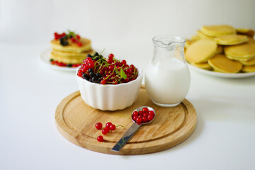 
Summer breakfast pancakes with red and black currants in a bowl, berries in a spoon, milk in a glass jug on a wooden board on a white background.