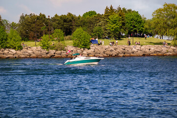 Fototapeta na wymiar MISSISSAUGA, CANADA - 06 10 2020: A green and white motorboat with Canadian flag gliding in the waters of Lake Ontario in front of people having rest at the Waterfront Trail