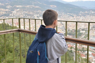 Little boy, looking at the amazing view on the Mediterranian sea gulf with touristic ships and aincient castle and a city from the mountain with a backpack on the back