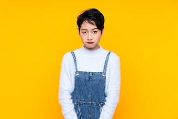 Young Asian girl in overalls over isolated yellow background sad