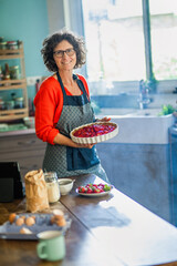 Portrait of a cheerful middle-aged woman in her kitchen. She proudly presents the strawberry tart she has just prepared.