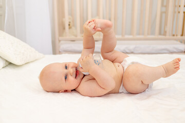 Fototapeta na wymiar baby boy 8 months old lying in diapers on a white bed with a bottle of milk at home feet up, baby food concept, baby drinking water from a bottle, place for text