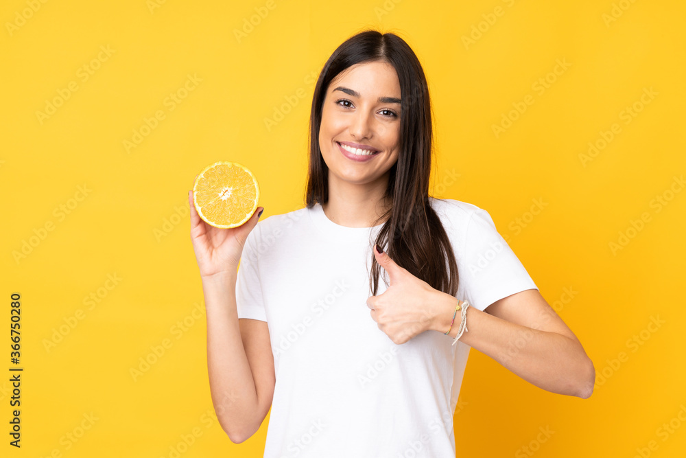 Wall mural Young caucasian woman holding an orange isolated on yellow background with thumbs up because something good has happened