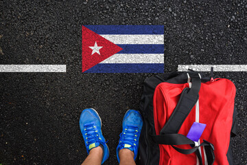 a man with a shoes and travel bag is standing on asphalt next to flag of Cuba and border
