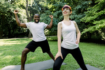 Multiethnic fitness people standing on a mat doing different exercises. Caucasian pretty woman with smile at workout in park outdoors. Focus on smiling African man keeps his fists up standing behind