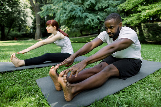 Image of smiling fitness multiethnic couple, wearing sports clothes, working out and stretching legs while sitting on exercise mats in green park outdoors