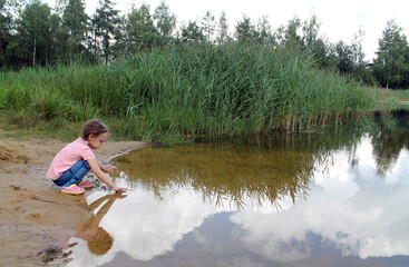 Fototapeta na wymiar A little girl plays by a pond against the background of green thickets. The concept of a happy childhood and outdoor recreation