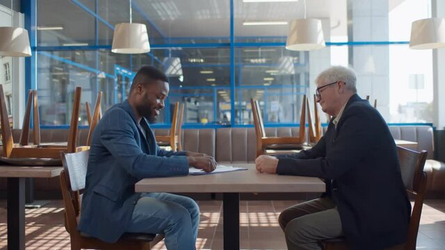 Side view of young african entrepreneur mature bank manager signing contract in empty cafe
