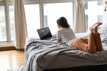 Fototapeta premium A beautiful young brunette girl is working on the laptop while lying on a bed by a panoramic window. Stylish modern interior. A cozy workplace. Shopping on the Internet.