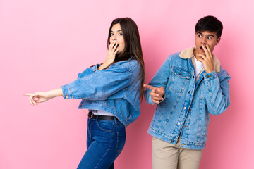 Young couple over isolated pink background pointing finger to the side with a surprised face