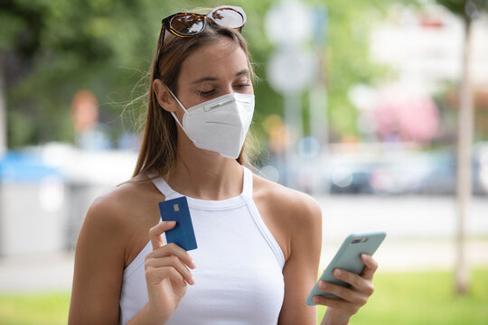 Happy Woman Paying Online With Credit Card On Smart Phone Avoiding Coronavirus With Mask Outdoors