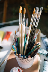 White pot with new paint brushes and pencils. Close-up view of new and used brushes in white pen pot with colorful pencils.