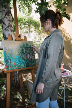 Side view of girl painting picture outdoor. Back of brunette woman painting on canvas placed on tripod in backyard with plants and flowers.