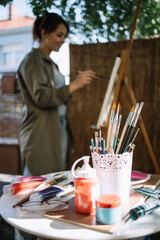 Table with paint tubes and brushes in front of blurred artist. Close-up view of table with painting accessories near out of focus female artist painting on canvas.