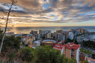 Spanish coast in Andalusia in the morning. View over the city of Malaga at sunrise with clouds and an orange horizon. City view on the Costa del Sol from park with trees and bushes