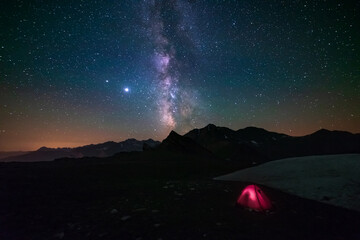 Milky way galaxy stars in the night sky over the Alps, illuminated camping tent in foreground,  snowcapped mountain range, astro photography stargazing