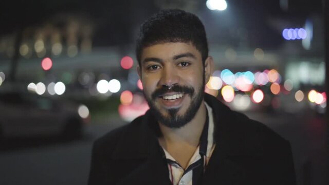 Attractive bearded young Arabic man in overcoat standing on city street, using gadget, looking down on mobile phone, then turning face to camera and smiling. Night, closeup shot. Male portrait concept