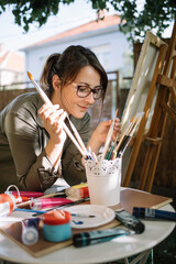 Woman wearing glasses and choosing paint brush from pot. Young woman taking paint brush from white pot with brushes in backyard.