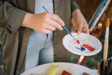 Woman's hands mixing paints in plate using brush. Cropped artist mixing paints and making palette while standing next to table with brushes and pencils.