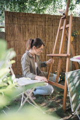 Girl sitting in garden with wooden stand and painting. Young woman squatting in garden while...
