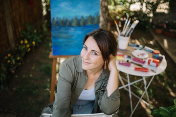 Portrait of woman painter posing in garden art studio. High angle view of woman artist sitting in outdoor art studio and looking at camera.