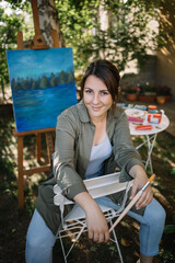 Young attractive woman artist sitting in outdoor art studio. Female artist sitting on chair in front of painted canvas and table with paint tubes in yard.