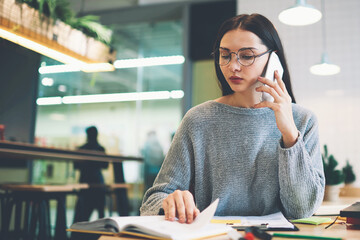 Concentrated attractive young student preparing homework reading textbook about media marketing while creating business plan consulting with classmate project partner through phone conversation