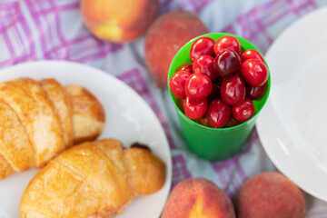 Picnic lunch food outdoors in summer in the Park. Purple, pink flowers in a basket, peaches, croissants, berries on a tablecloth in nature