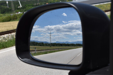 Landschaft mit Strommasten, Himmel, Wolken und Straße, Eisenbahnschienen im Rückspiegel, Blick in den Rückspiegel eines Autos