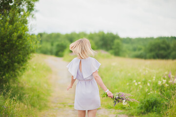 Fototapeta na wymiar A young girl in a purple dress with a bouquet of wild flowers in her hands is spinning on the road in a field in summer. A girl enjoys a walk in the Park.