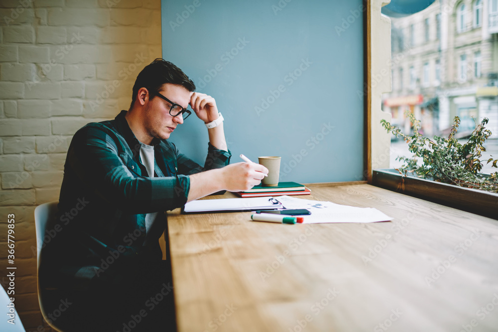 Poster pensive hipster guy studying in coworking space learning information from notings and textbooks for 