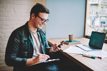 Cheerful young man laughing on funny video browsed on smartphone while working remotely in cafe...