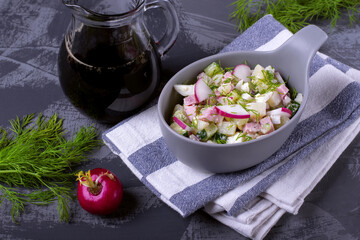 Okroshka soup with radish, sausage, cucumber, egg and potato in a ceramic bowl. Kvass in glass jug on the grey table