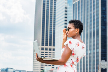 Young African American Woman traveling, working in New York City, with short afro hair, wearing dress, sunglasses, standing in front of business district, working on laptop computer, looking, thinking