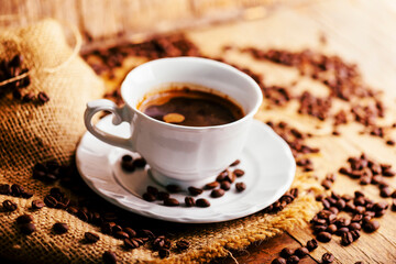 Closeup of coffee cup and coffee beans with vintage burlap on rustic wooden table. Hot beverage in mug still life.