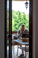 Portrait of handsome successful man using smartphone while looking at laptop.Shot of male working on the balcony at home.Young attractive man sitting and doing some work on laptop and using mobile.