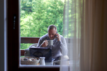 Shot of young man using smartphone and having coffee on the sofa,while looking at laptop.Young serious caucasian bearded businessman dressed casual using mobile and drinking coffee.Technology concept.