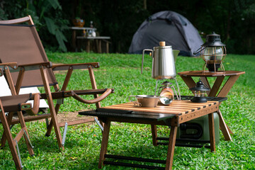 Stainless steel kettle, portable gas stove, bowl and vintage lanterns with outdoors table set on green lawn in camping area