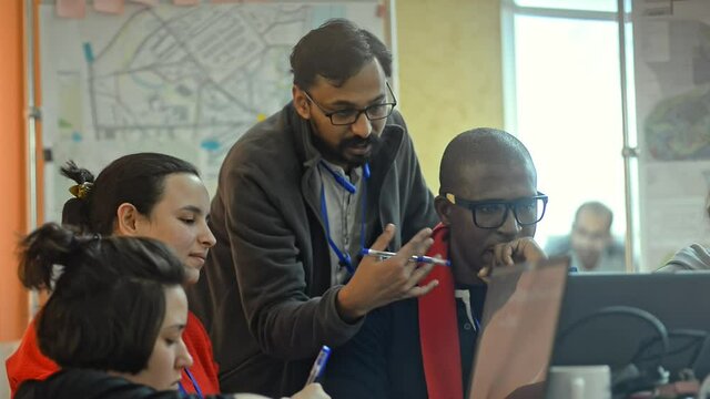 Young multi-ethnic people working on online project together at table in modern office irrl. Cheerful men and women are talking about startup and sharing ideas at desk with laptops. Multiracial team