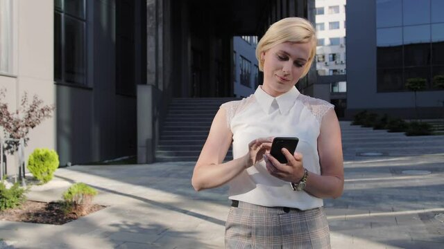 Beautiful and confident blonde in a white shirt installs the application on a mobile phone outdoor or chats with colleagues.