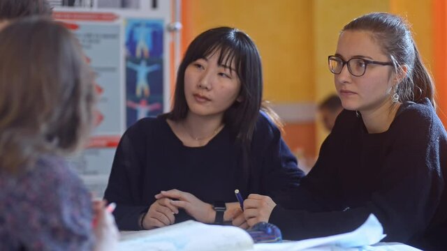 Multiethnic students study together while sitting at desk in university irrl. Young cheerful girls and guy talking with smiles and doing paper work at table in room. Millennial multiracial team is in