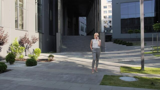 Attractive blonde in a white shirt and a beautiful watch speaks on a mobile phone and smiles outdoors on the background of a large glass building.Blonde Girl. Successful Woman.