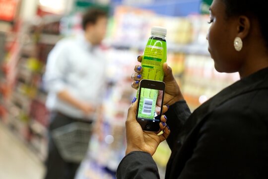 Employee Photographing Soft Drinks In A Supermarket Aisle With A Customer Shopping In The Background.