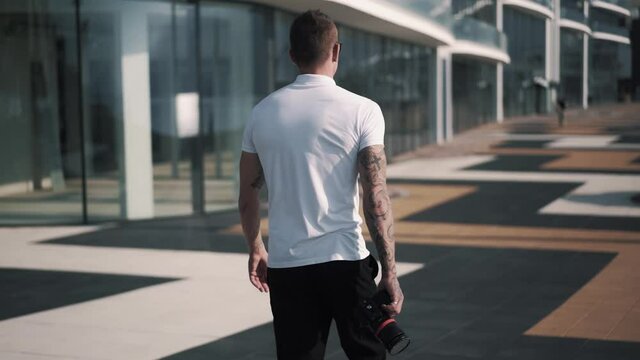 Backside view of tattooed male photographer walking with camera along street