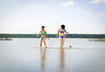 Two teenage sisters in the heat of the lake run with splashes on the water with a dog to the shore.
