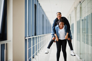 Two african woman friends in jeans jacket having fun, jumped on shoulders indoor together.