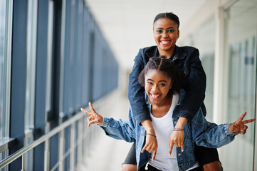 Two african woman friends in jeans jacket having fun, jumped on shoulders indoor together.