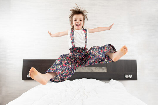 Content kid in overalls in moment of jumping on bed with outstretched arms and legs looking at camera