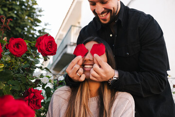 Low angle of cheerful young man covering eyes of laughing girlfriend with petals of red rose while having fun and enjoying romantic date in city